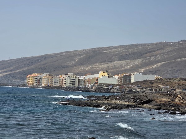 Blick entlang der Küste auf die Häuserfront von Puertito de Güímar; davor zerklüftete Lavafelsen und wogende Wellen.
