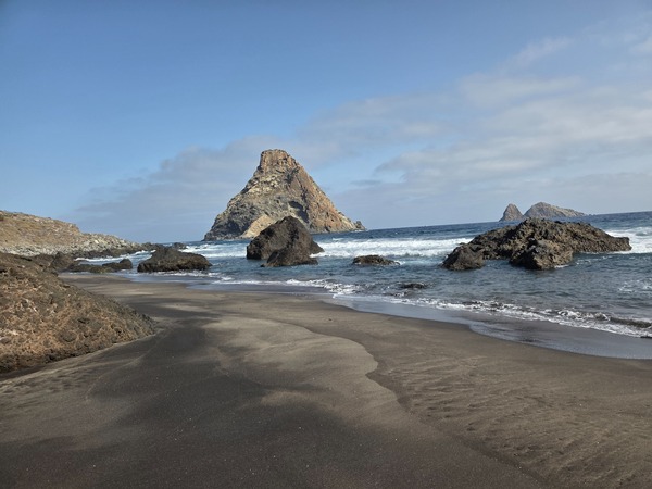 Schwarzer Sandstrand der Playa de Las Breñas mit markantem Felsen und kleinen Riffen vor der Küste, Wellen rollen in die Bucht.