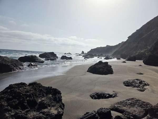 Weite Sandfläche mit verstreuten Basaltbrocken an der Playa de Las Breñas, dahinter die steilen Hänge des Anaga-Gebirges im Gegenlicht.