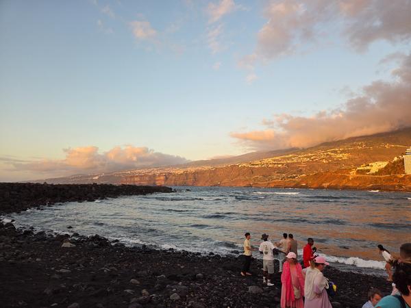 Menschen stehen am schwarzen Vulkansandstrand von Puerto de la Cruz bei Sonnenuntergang mit Blick auf die Küste des Orotava-Tals.