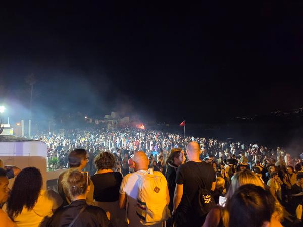 Große Menschenmenge am Strand bei Nacht, umgeben von Lagerfeuern und Musik.