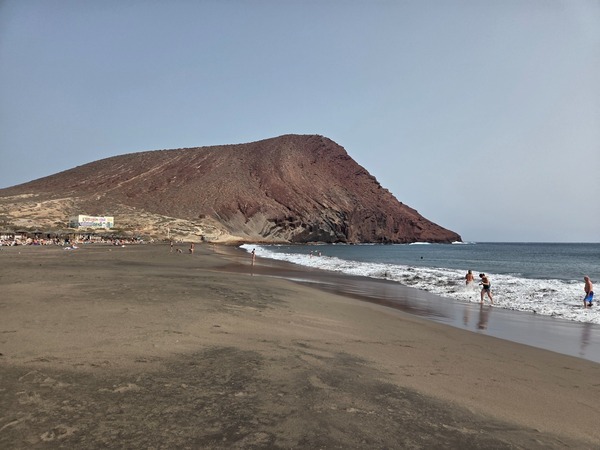 Breiter Strandabschnitt der Playa de La Tejita mit der rötlichen Montaña Roja im Hintergrund und Badenden am Ufer.