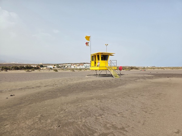 Gelber Rettungsturm an der Playa de La Tejita mit gehissten Warnflaggen vor dem Hintergrund von Dünen und Häusern.